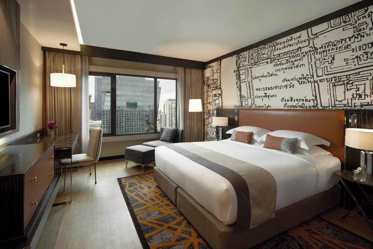 A hotel room with a large bed, a desk with a chair, a TV, and a view of the cityscape through large windows.