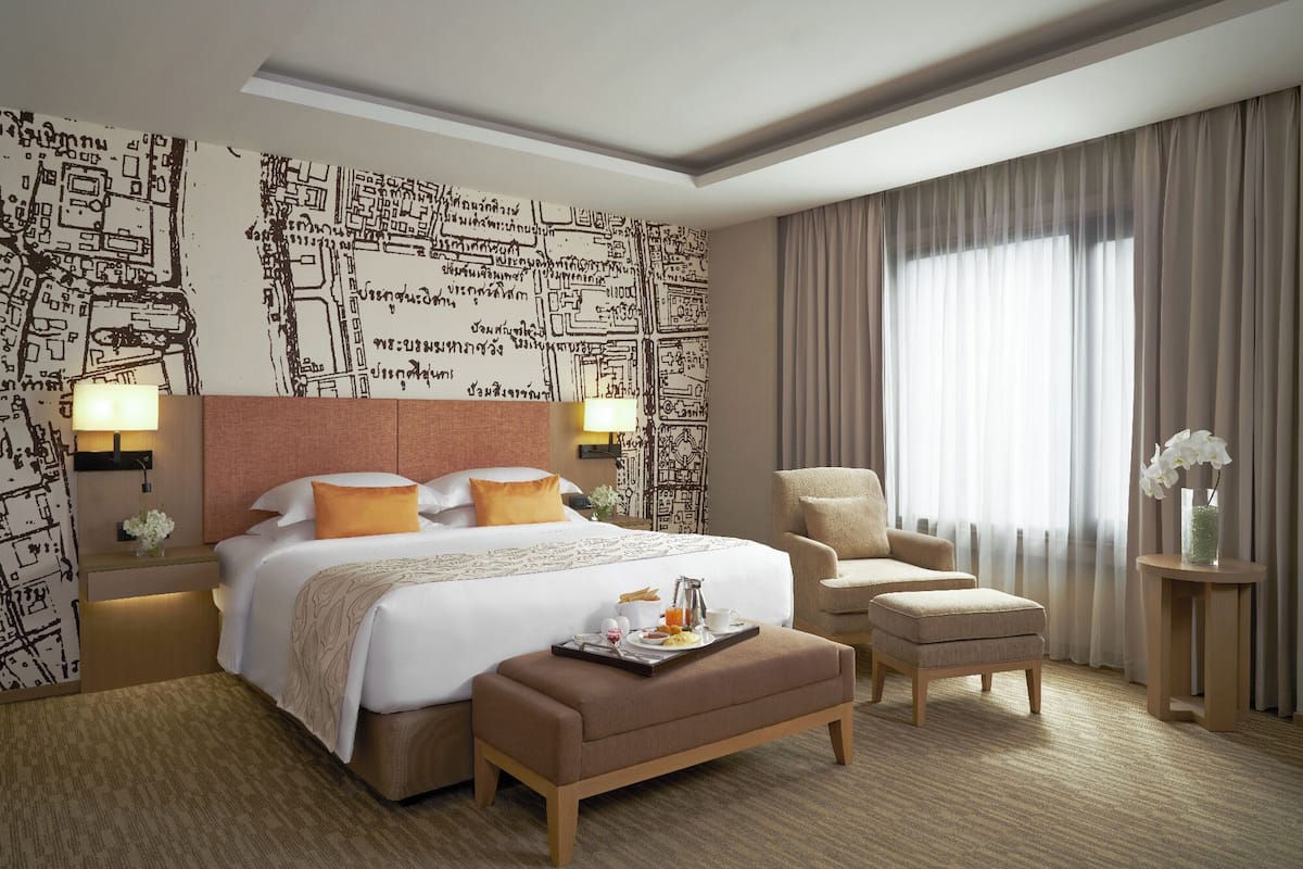A hotel room with a large bed, a chair, an ottoman, and a nightstand with a lamp.
