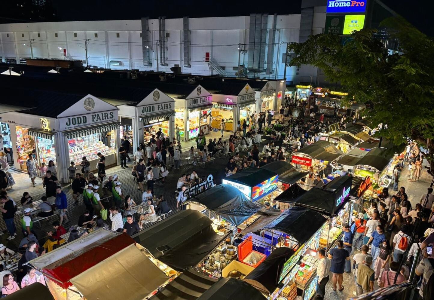 Um mercado ao ar livre movimentado com diversos quiosques e lojas, incluindo Jodd Fairs, sob um céu noturno.