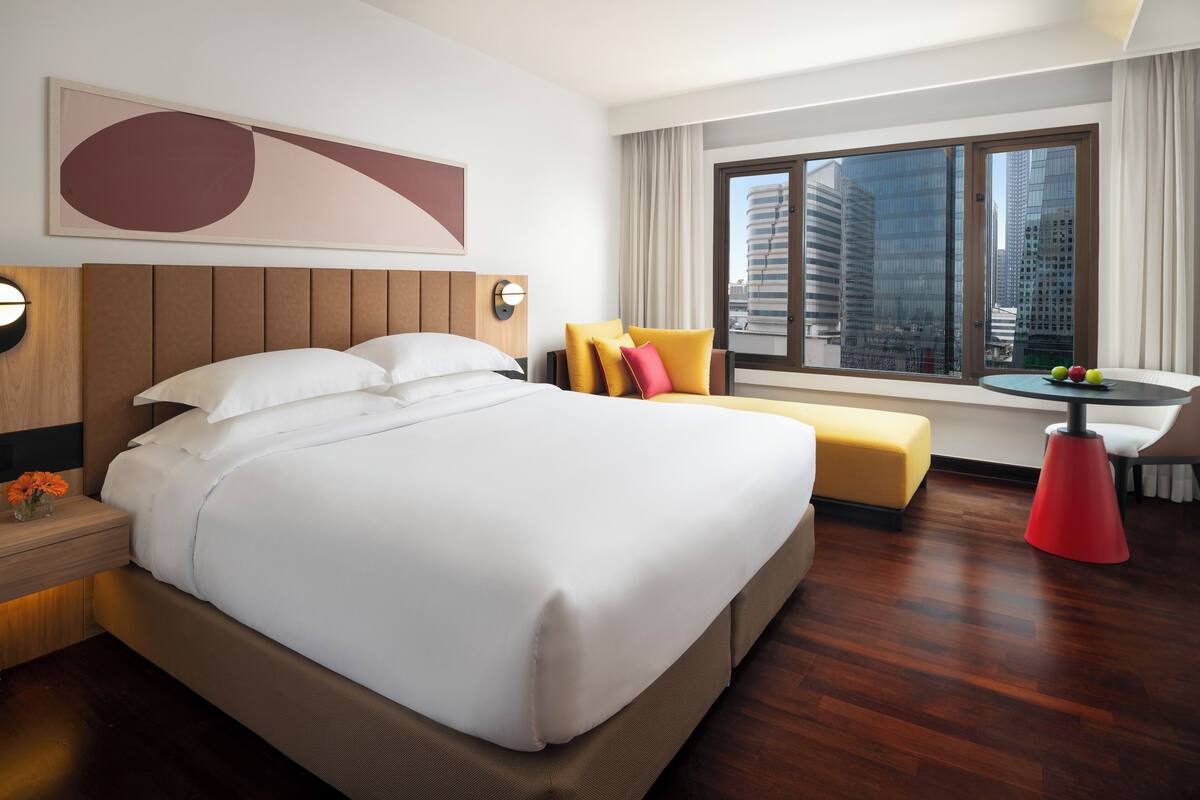 A hotel room with a large bed, a small table with fruit, a sofa, and a view of tall buildings.