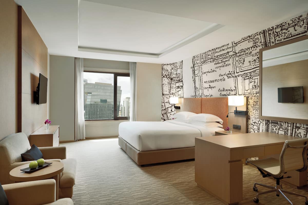 A hotel room with a bed, a desk, a chair, a small table with apples, a TV, and a window with a city view.