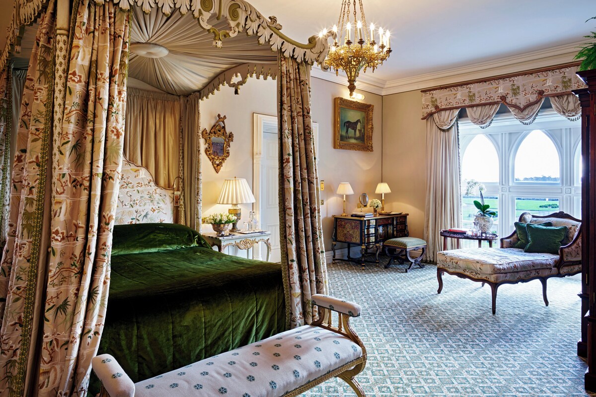 A luxurious bedroom with a four-poster bed, ornate curtains, a chandelier, and a view of the outdoors through arched windows.