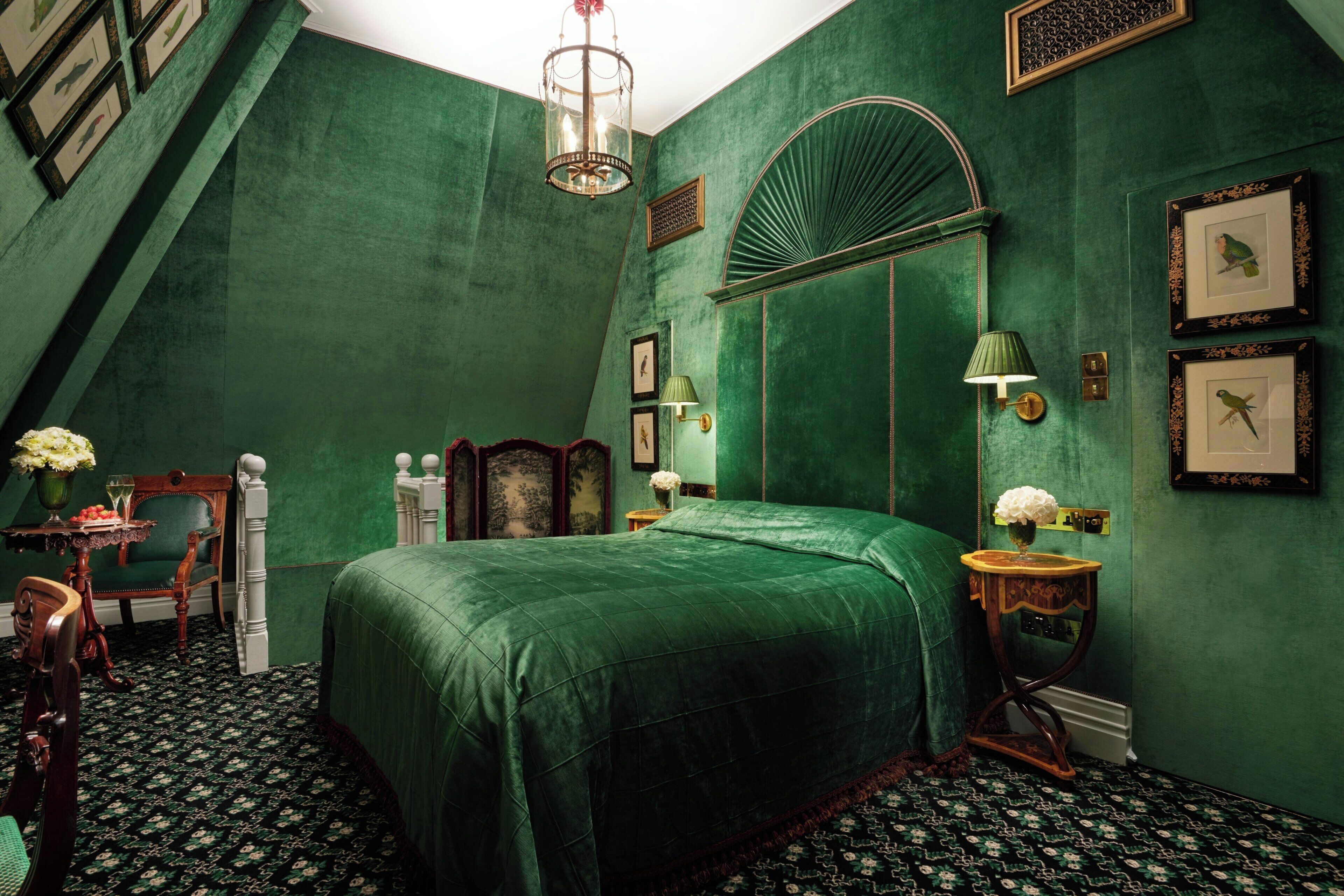 A room with a bed, a chair, a table with flowers, a lamp, and framed pictures on the wall.