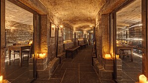 Un couloir aux murs de pierre, agrémenté de bancs et de tables, de bougies et de tableaux encadrés.