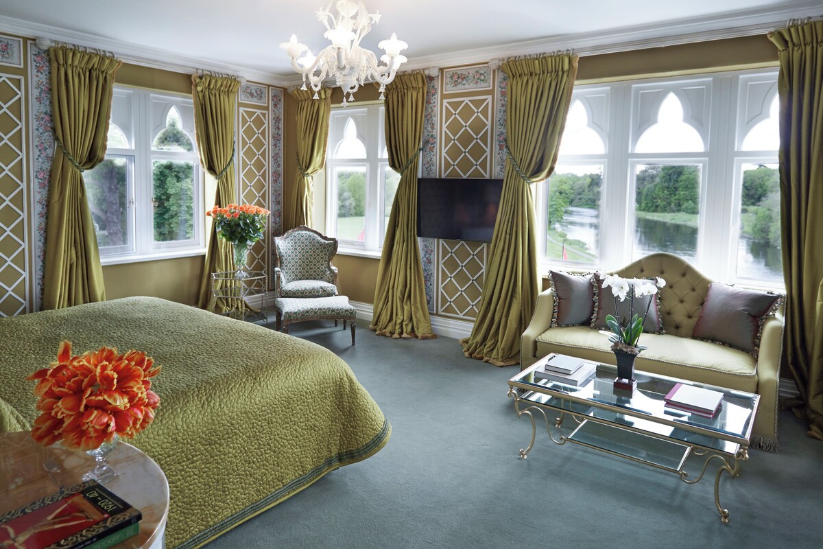 A spacious room with a large bed, a sofa, a chair, and a coffee table. There is a TV mounted on the wall and a window with curtains.