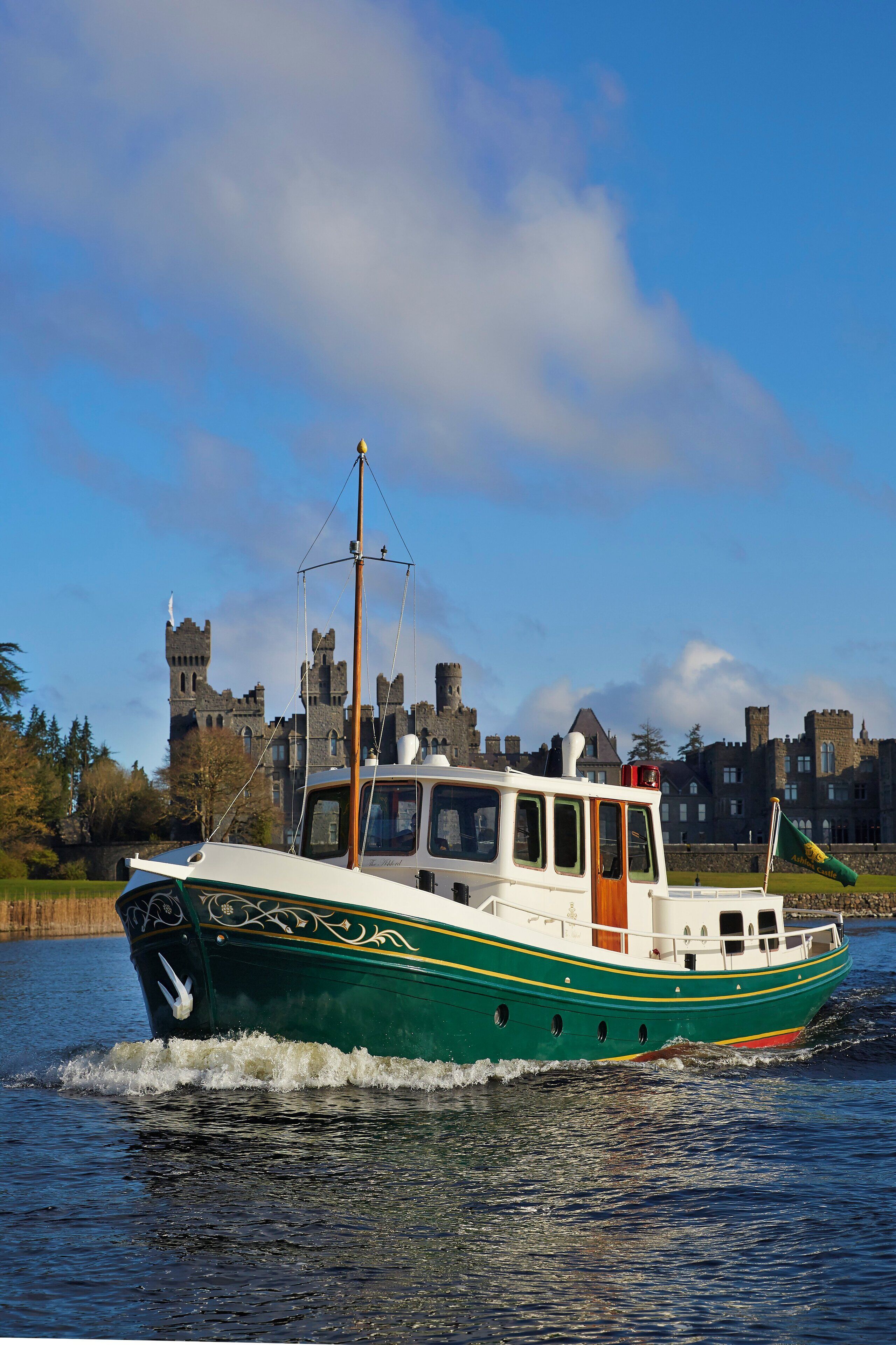 A green and white boat with a cabin, moving on water with a castle in the background.