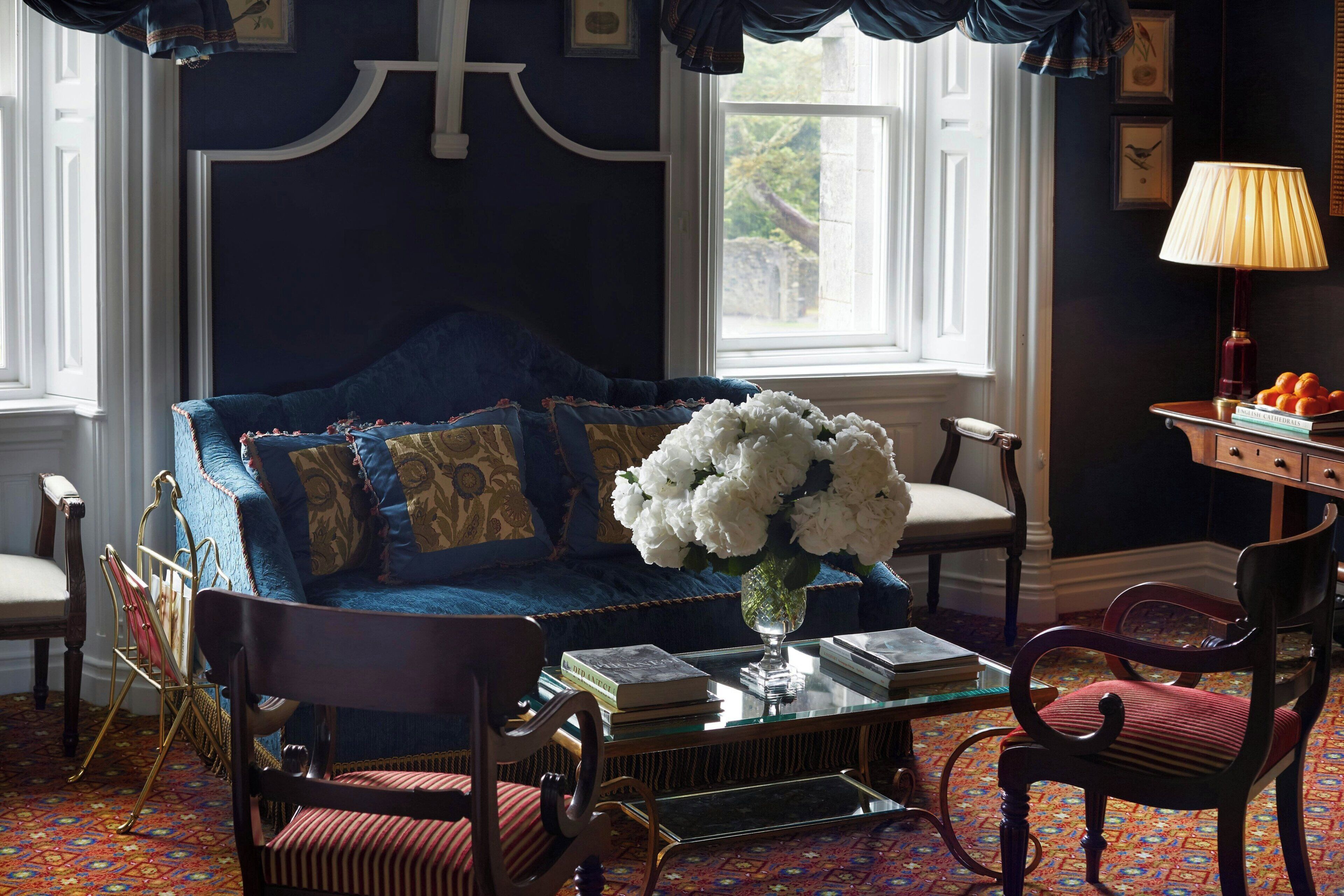 A room with a blue sofa, a glass coffee table with books and a vase of flowers, a wooden chair, and a lamp on a side table.
