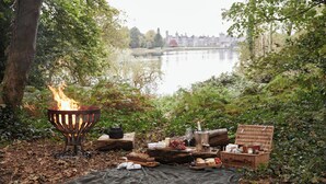 A campfire with a picnic setup by a lakeside, including a basket, food, and a bottle.