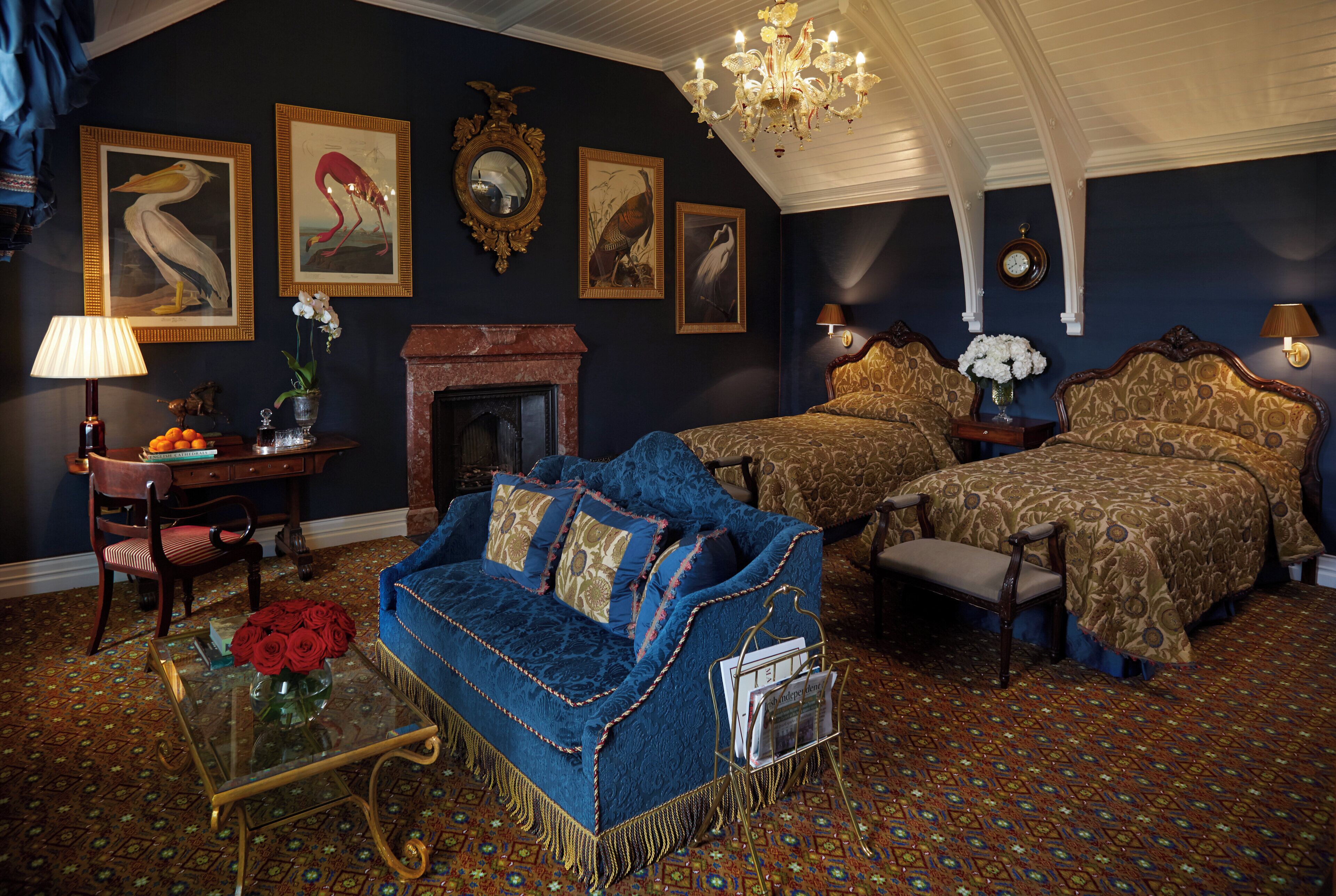 A room with a blue sofa, a fireplace, two beds with patterned bedding, a chandelier, and framed wall art.