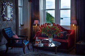 A room with a red sofa, a blue armchair, a glass coffee table with a vase of red flowers, and a view of the sea through large windows.