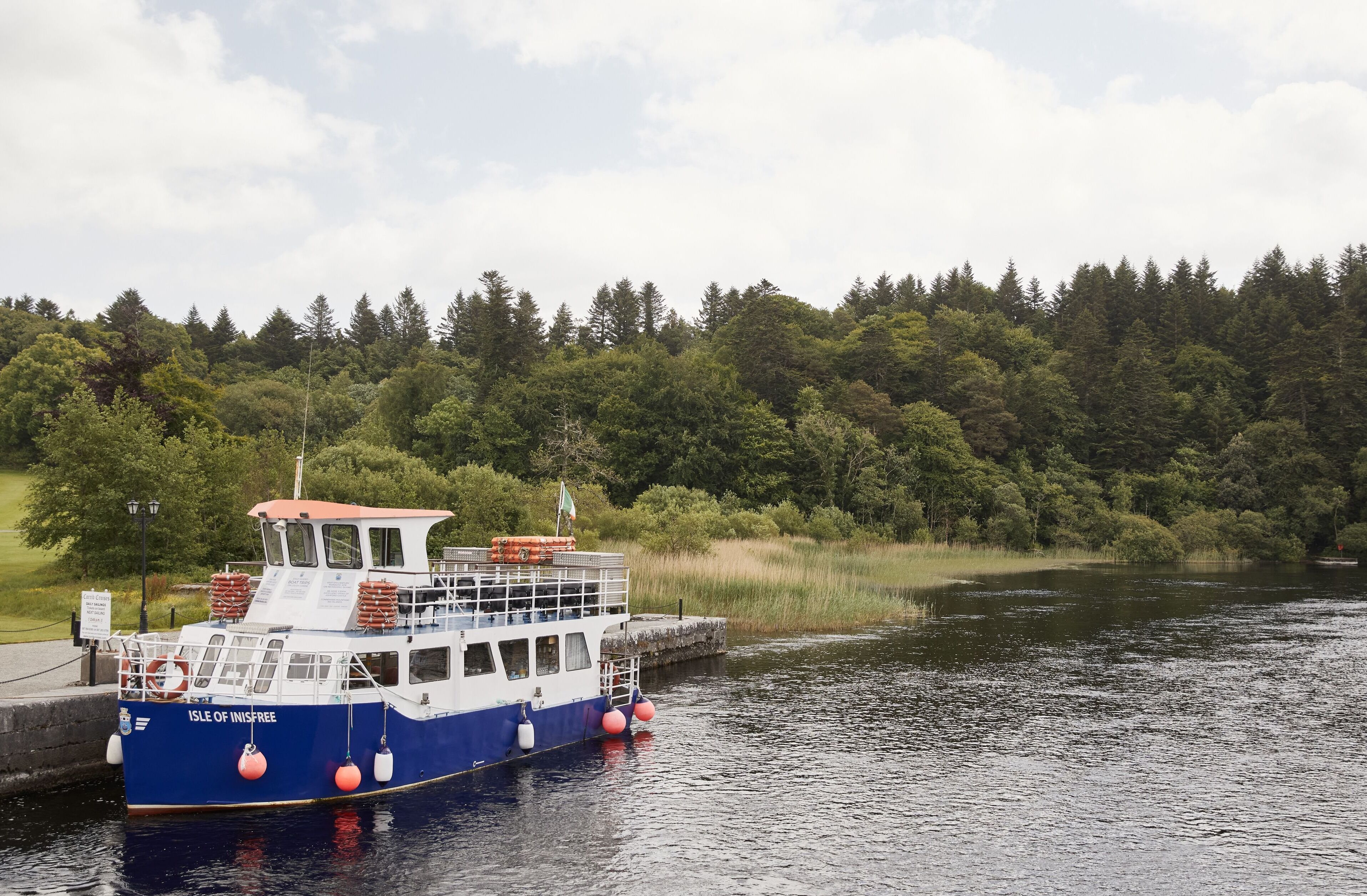 A boat named "Isle of Inisfree" docked at a pier with a forested shoreline in the background.