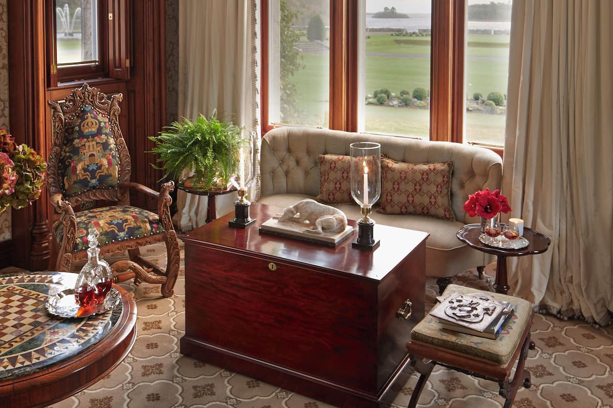 Una habitación con un ventanal grande, un sofá, un aparador de madera, una mesa redonda con un florero y una silla con cojín estampado.