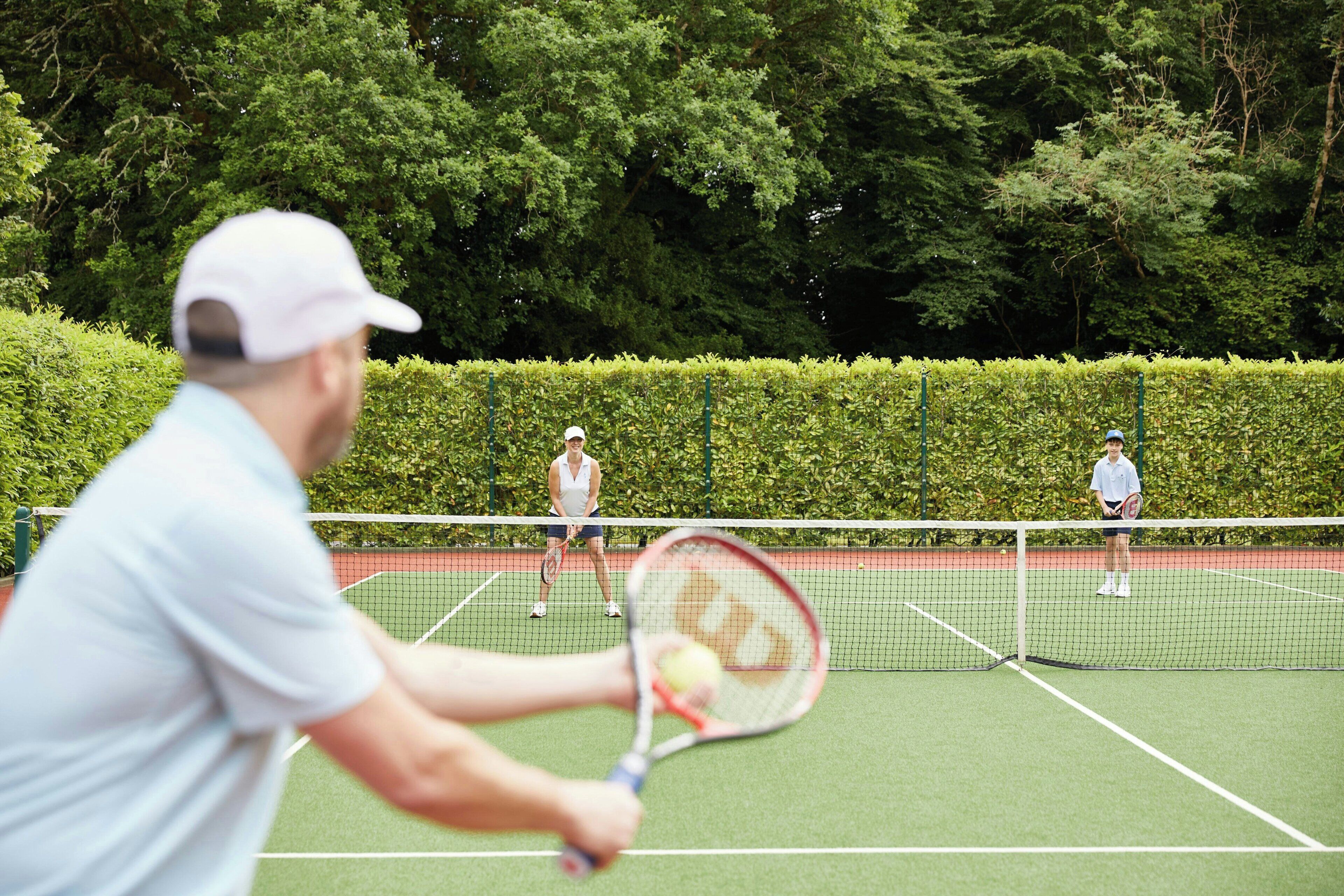 A tennis court with a net, surrounded by green hedges.