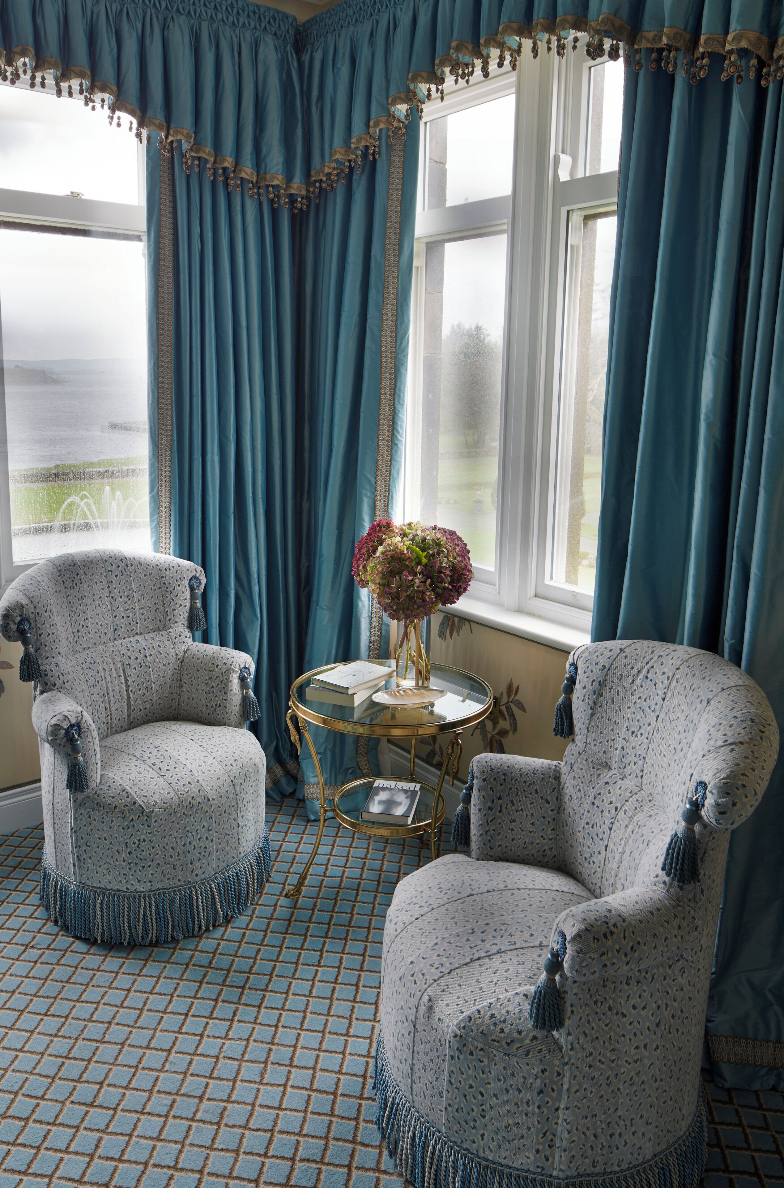 A room with two patterned armchairs, a small round table with books, and a vase of flowers.