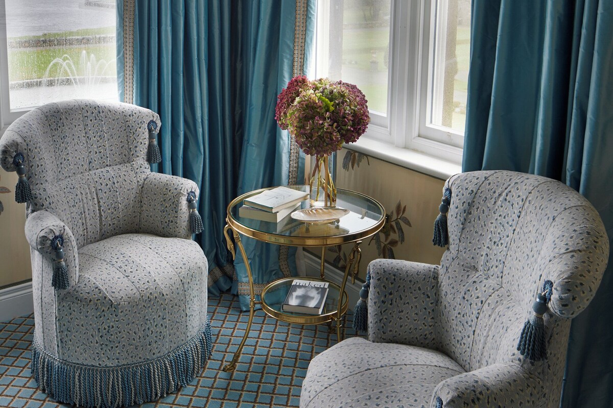 A room with two patterned armchairs, a small round table with books, and a vase of flowers.