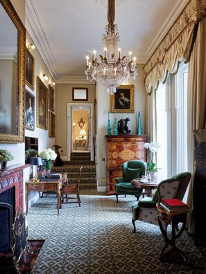 A grand room with a chandelier, green armchairs, a fireplace, and a staircase leading to a hallway.