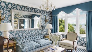 A room with a blue floral wallpaper, a tufted sofa, a white armchair, a chandelier, and a view of greenery through a window.
