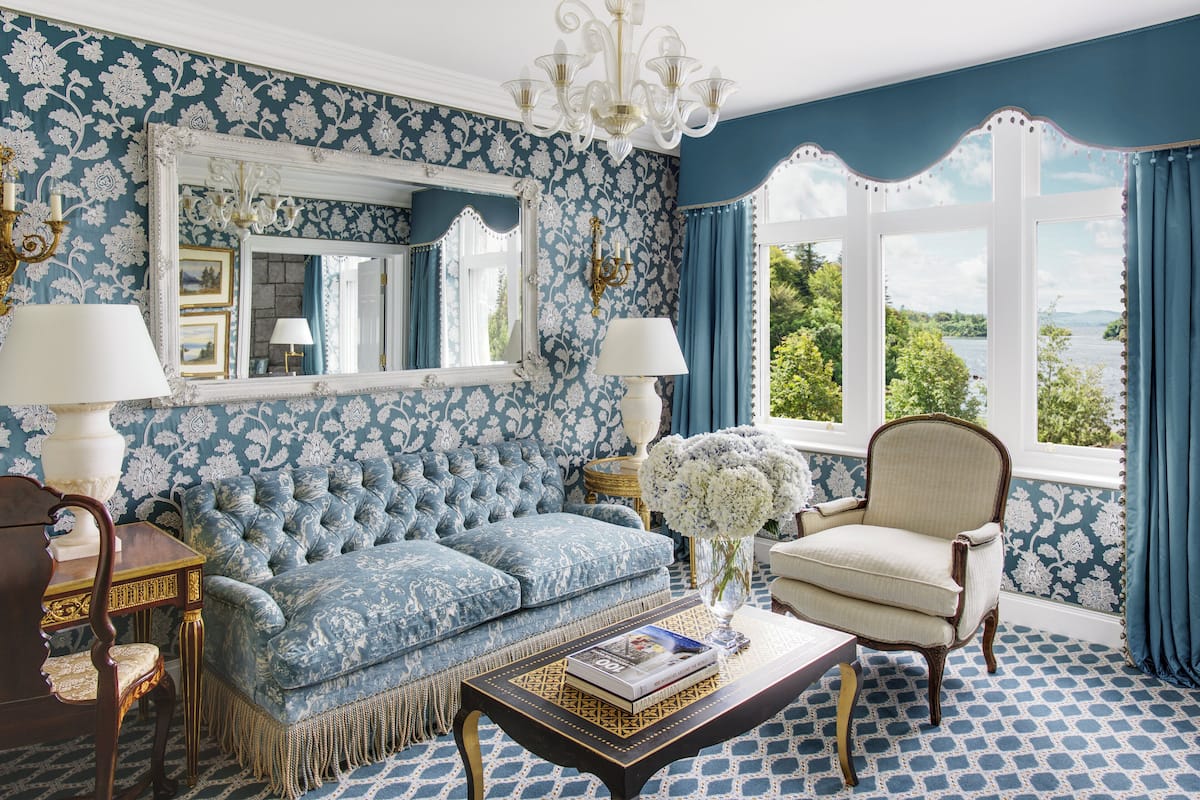 Una habitación con papel tapiz azul con motivos florales, un sofá capitonado, un sillón blanco, una araña y vistas al verdor a través de una ventana.