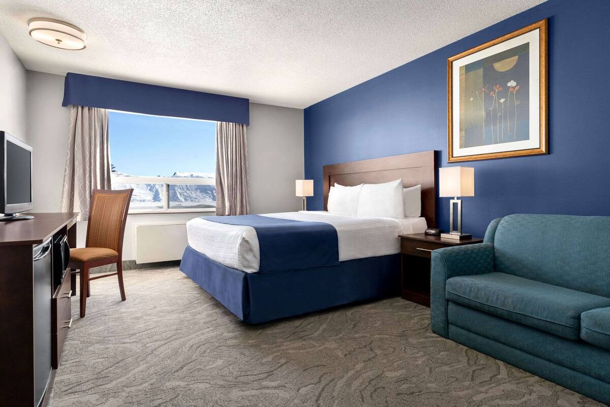 A hotel room with a bed, a desk, a chair, a sofa, a window with curtains, a painting on the wall, and a view of the mountains.