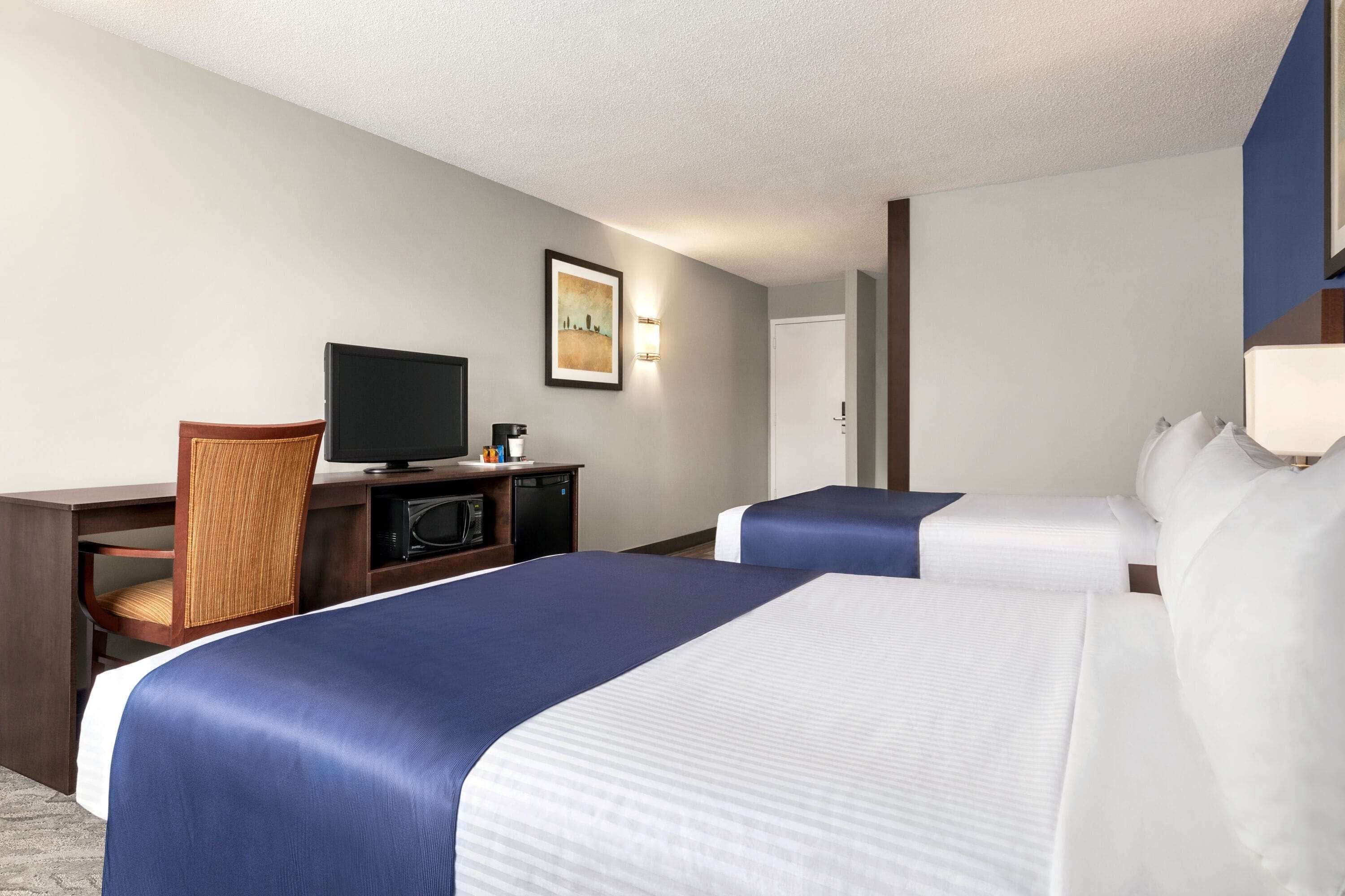 Ein Hotelzimmer mit zwei Betten, einem Schreibtisch, einem Fernseher, einer Mikrowelle und einem gerahmten Bild an der Wand.