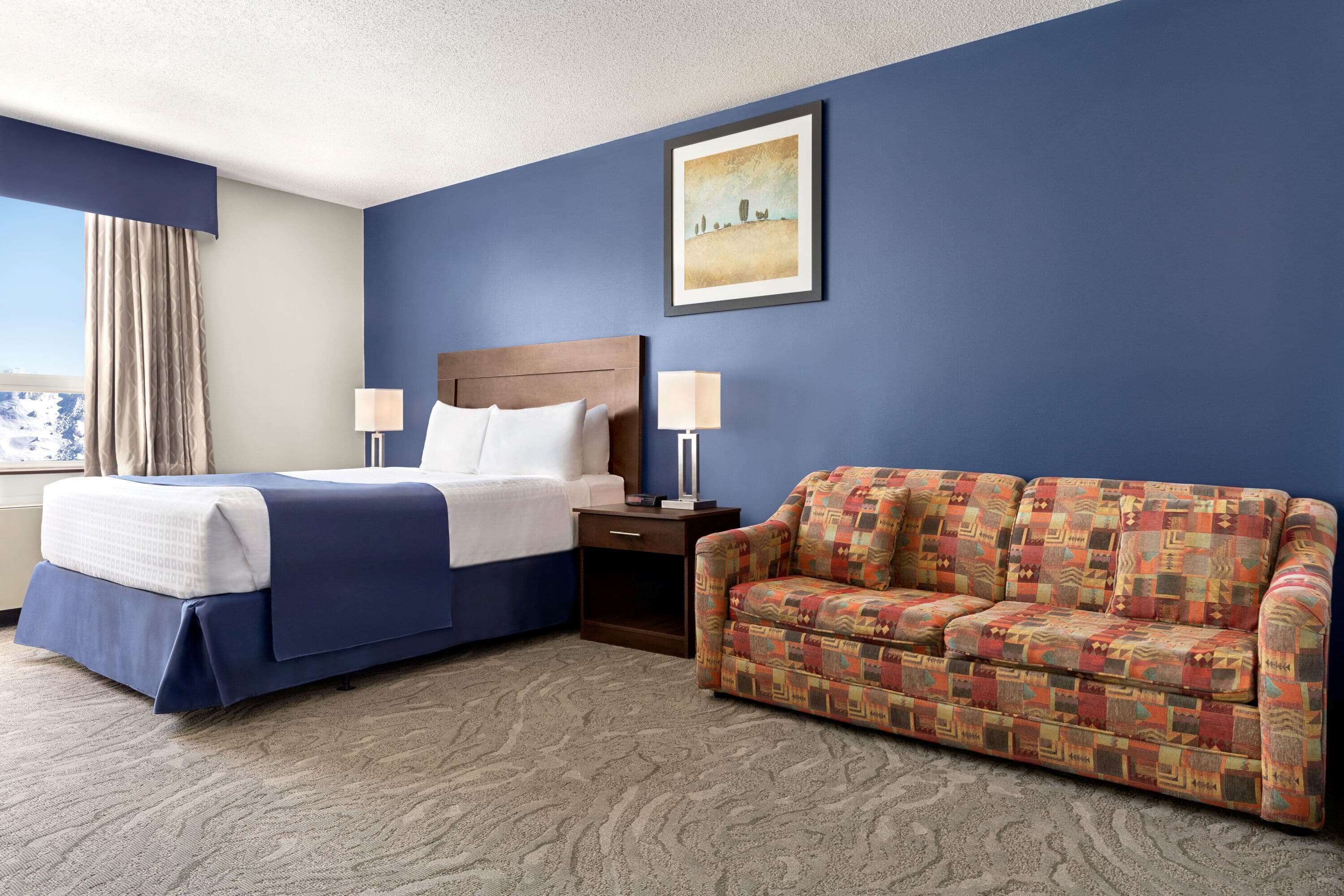 A hotel room with a bed, a sofa, a nightstand, a lamp, and a painting on the wall.