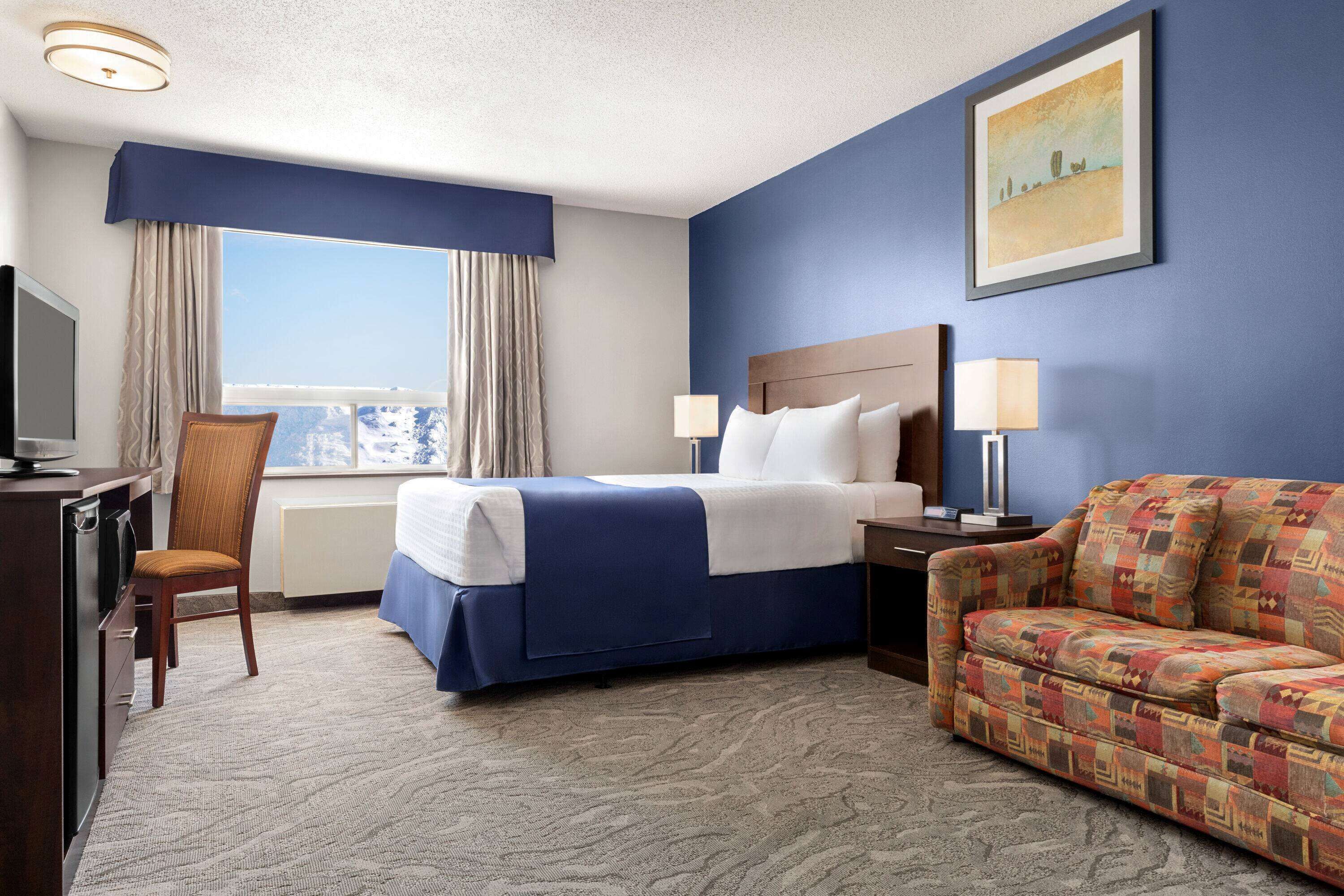 Habitación de hotel con cama, sofá, escritorio con televisor y ventana con cortinas.