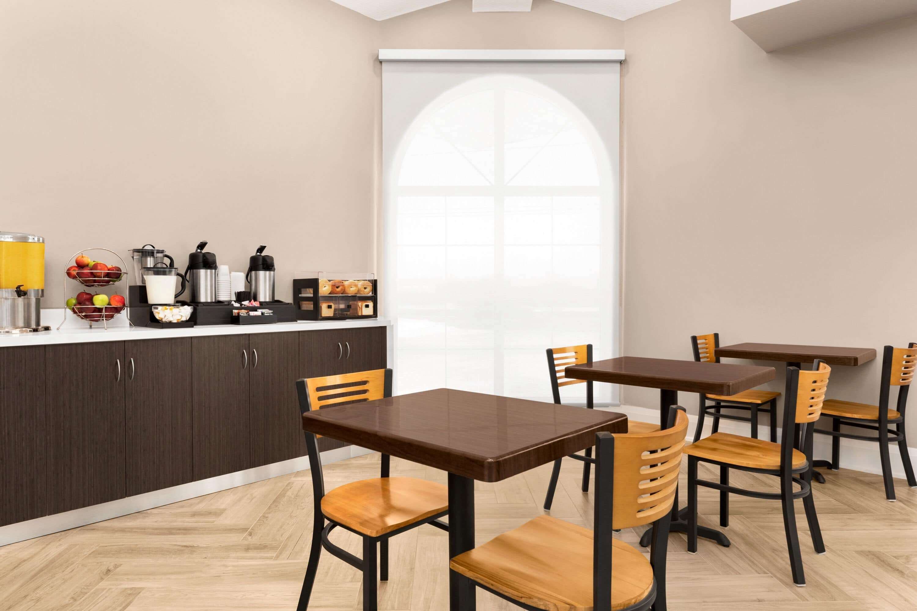 A modern dining area with wooden tables and chairs, a kitchen counter with appliances, and a large window with a view.