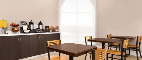 A modern dining area with wooden tables and chairs, a kitchen counter with appliances, and a large window with a view.