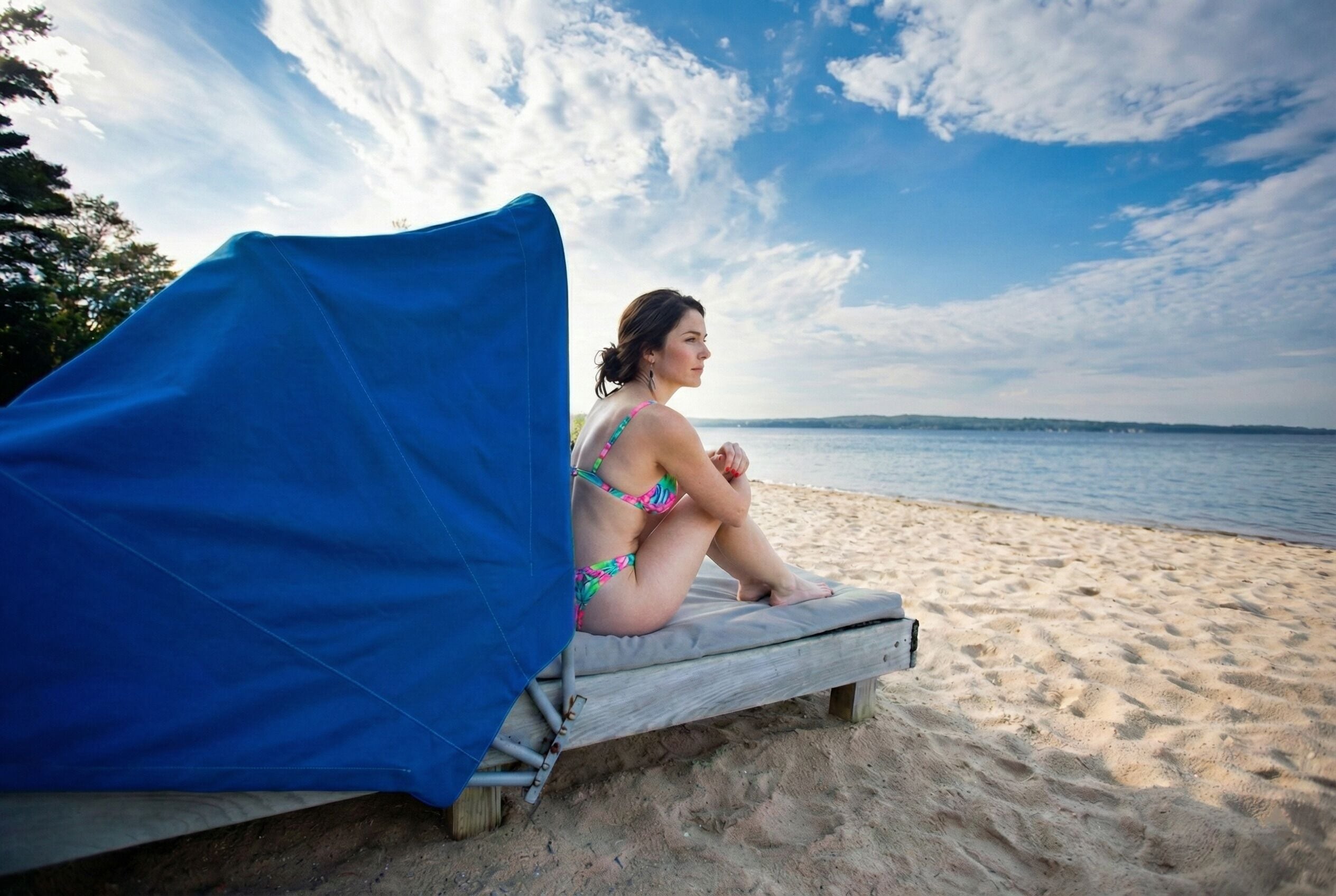 A person sitting on a wooden lounge chair with a blue cover, on a sandy beach with a view of the sea and a partly cloudy sky.