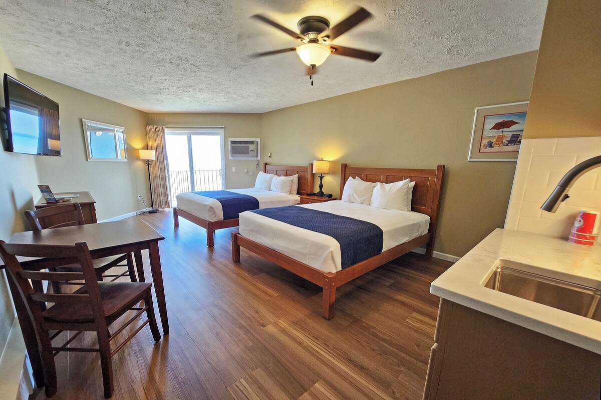 A hotel room with two beds, a desk, a chair, a ceiling fan, a window, and a kitchenette.