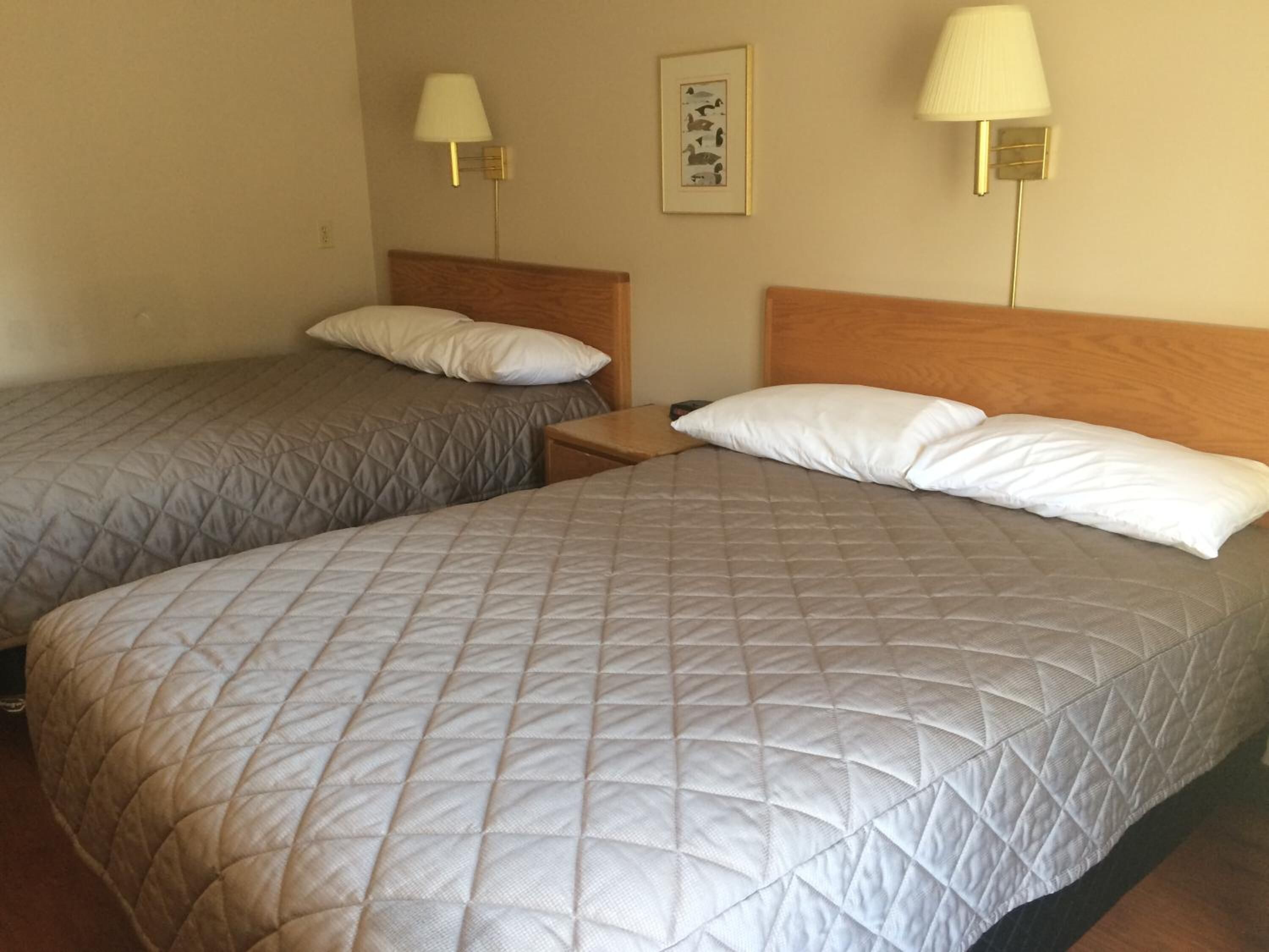 A hotel room with two beds, each with a quilted bedspread and a wooden headboard. There are two bedside lamps and a framed picture on the wall.