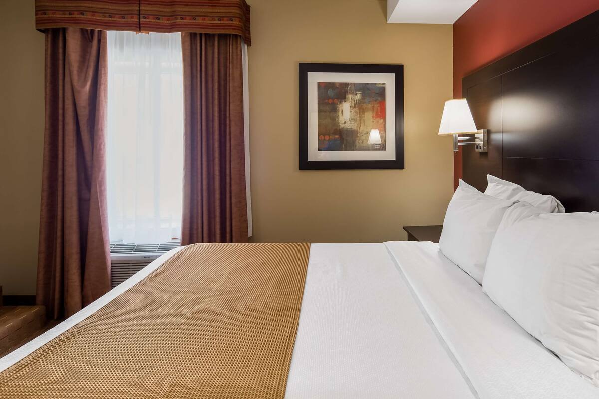 A hotel room with a large bed, a framed artwork on the wall, a bedside lamp, and a window with curtains.