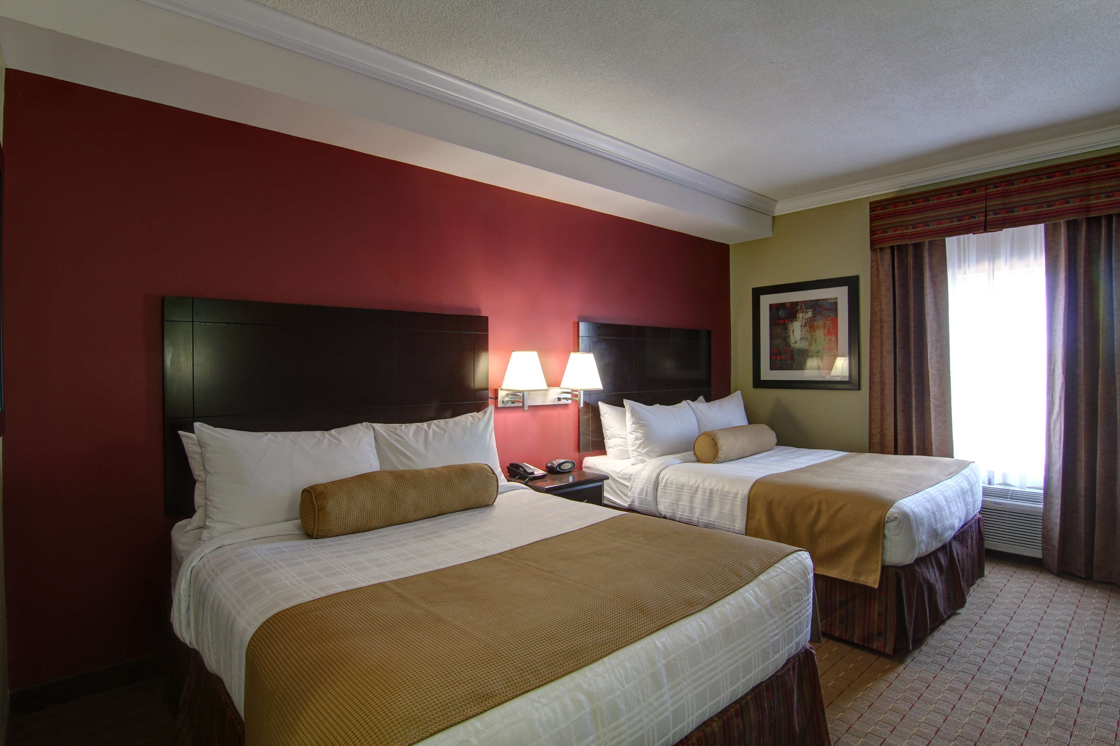 A hotel room with two beds, a red accent wall, a painting, and a window with curtains.