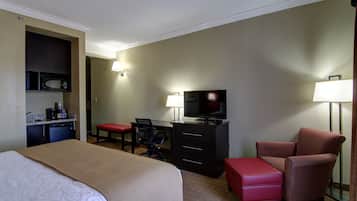 Una habitación de hotel con cama, escritorio con televisor, silla, reposapiés rojo y microondas en la kitchenette.