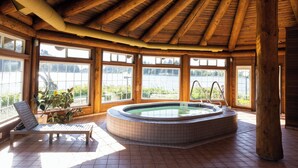 A wooden cabin interior with a hot tub, large windows, and a lounge chair.