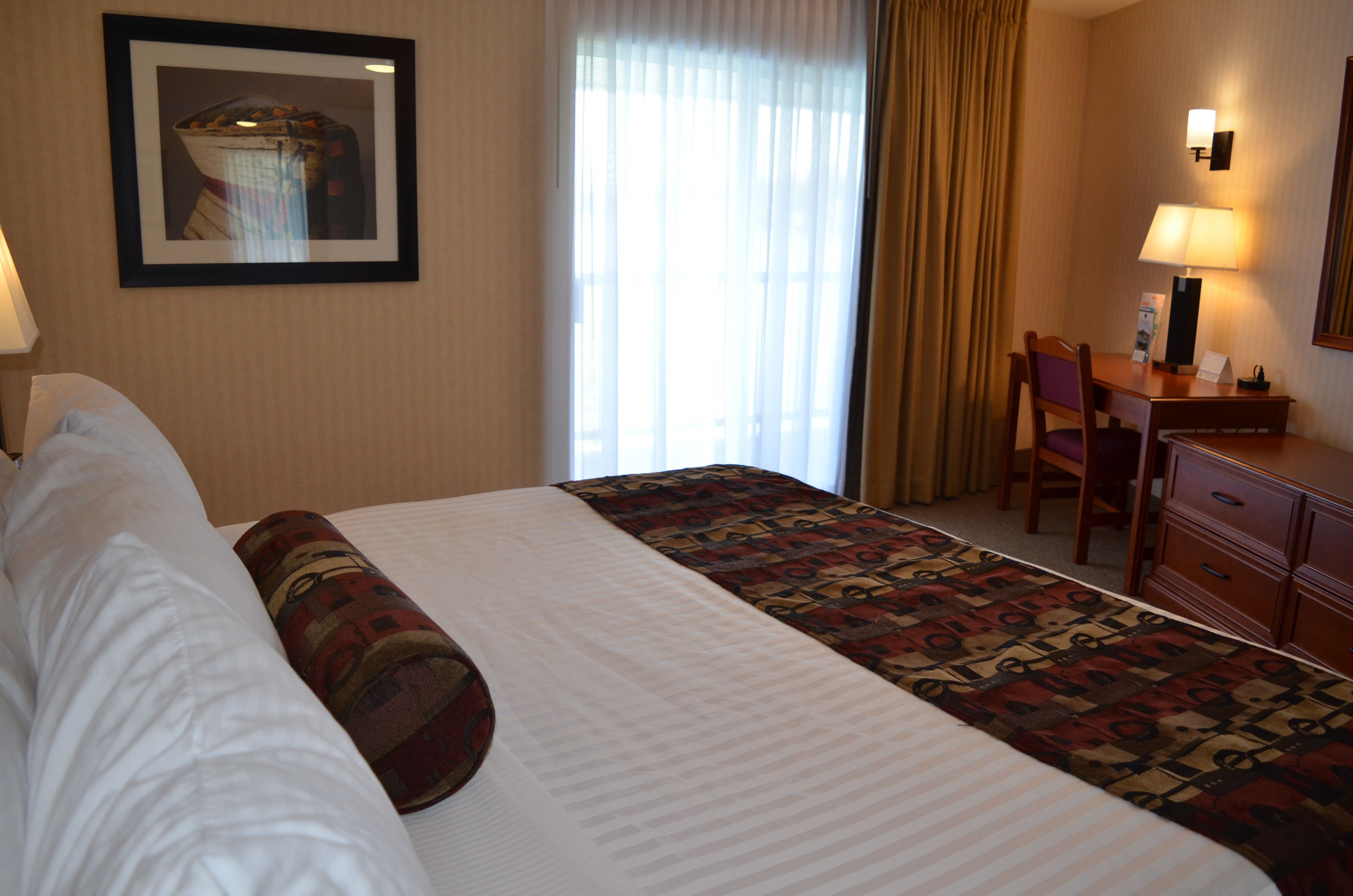 A hotel room with a bed, a desk, a chair, a lamp, and a framed picture on the wall.