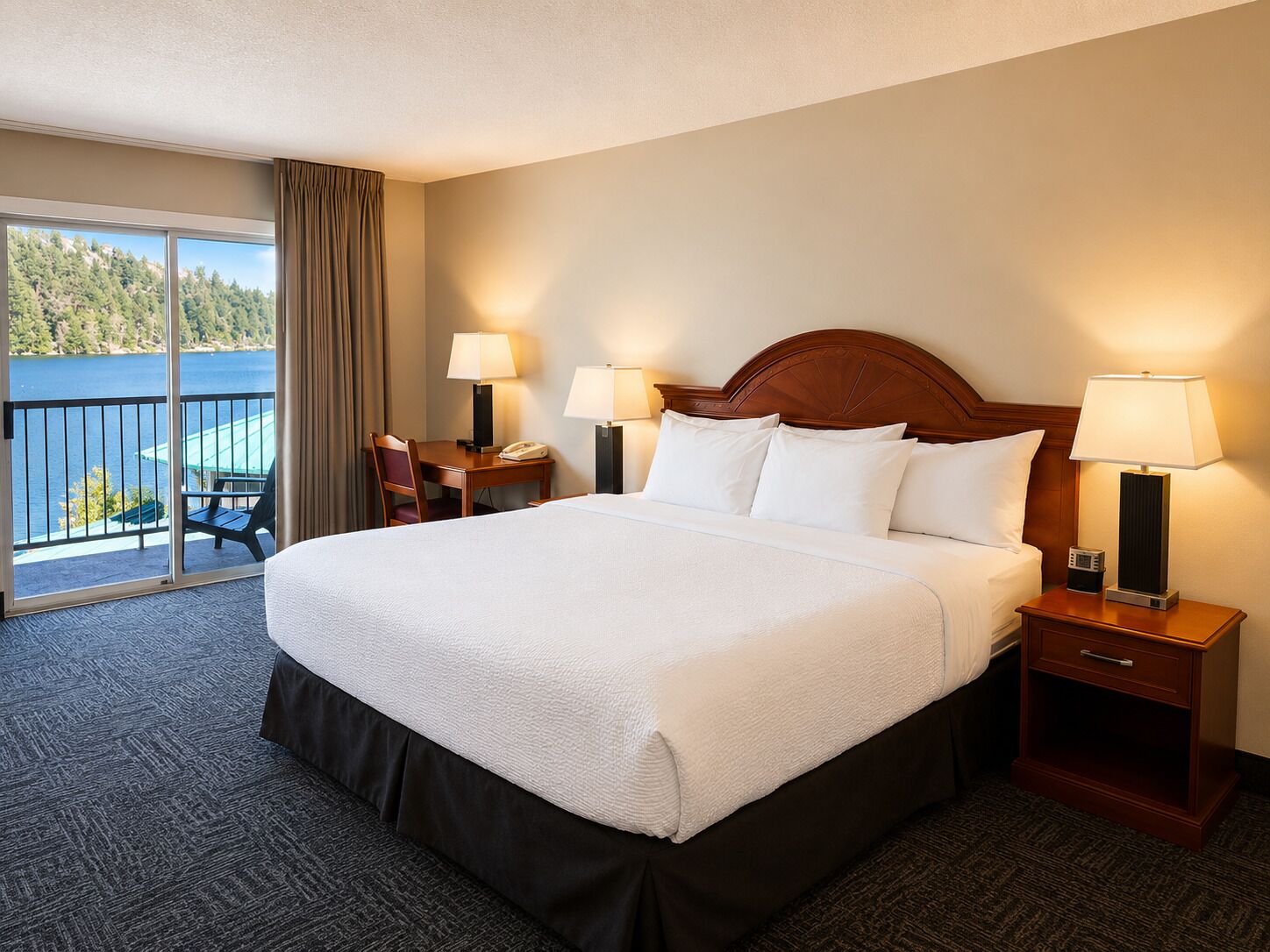 A hotel room with a large bed, two bedside tables with lamps, a desk with a chair, and a view of a lake through a sliding door.