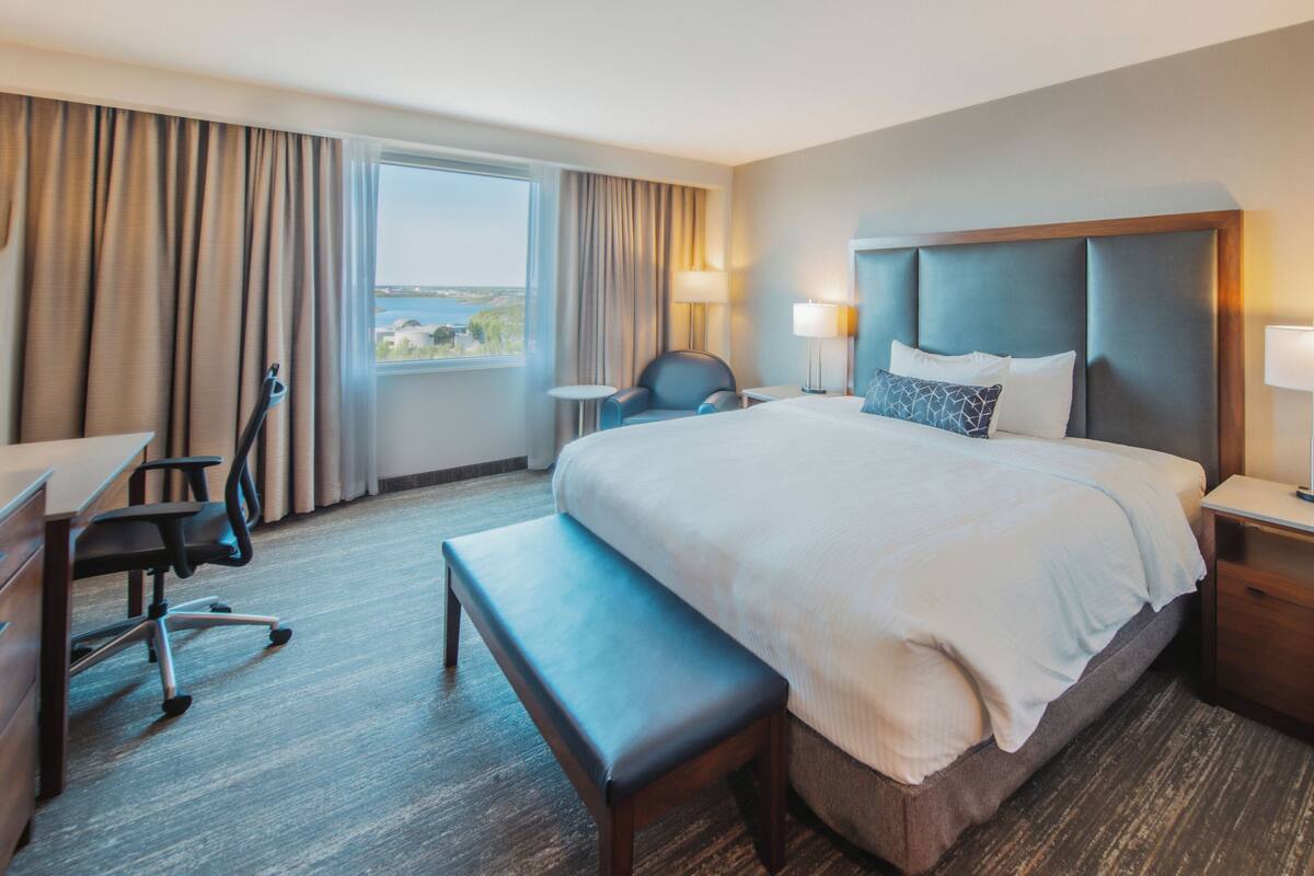 A hotel room with a large bed, a desk, a chair, a bench, and a view of the sea through the window.