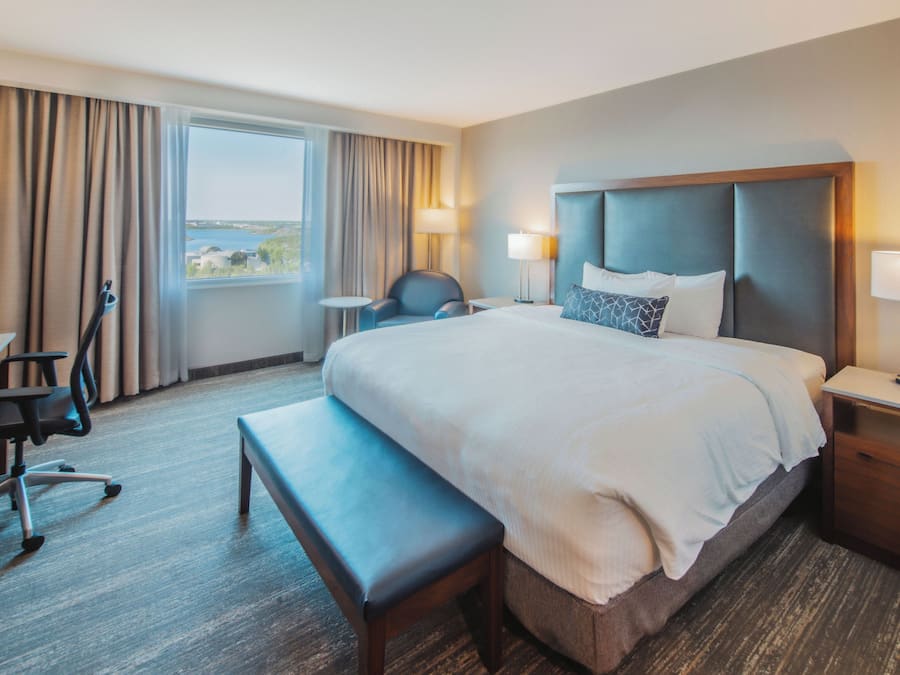 A hotel room with a large bed, a desk, a chair, a bench, and a view of the sea through the window.