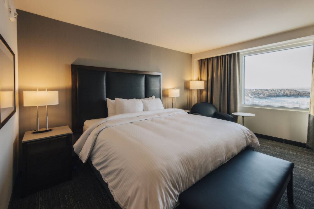 A hotel room with a large bed, two bedside tables with lamps, a small round table, a bench, and a view of a snowy landscape through a window.