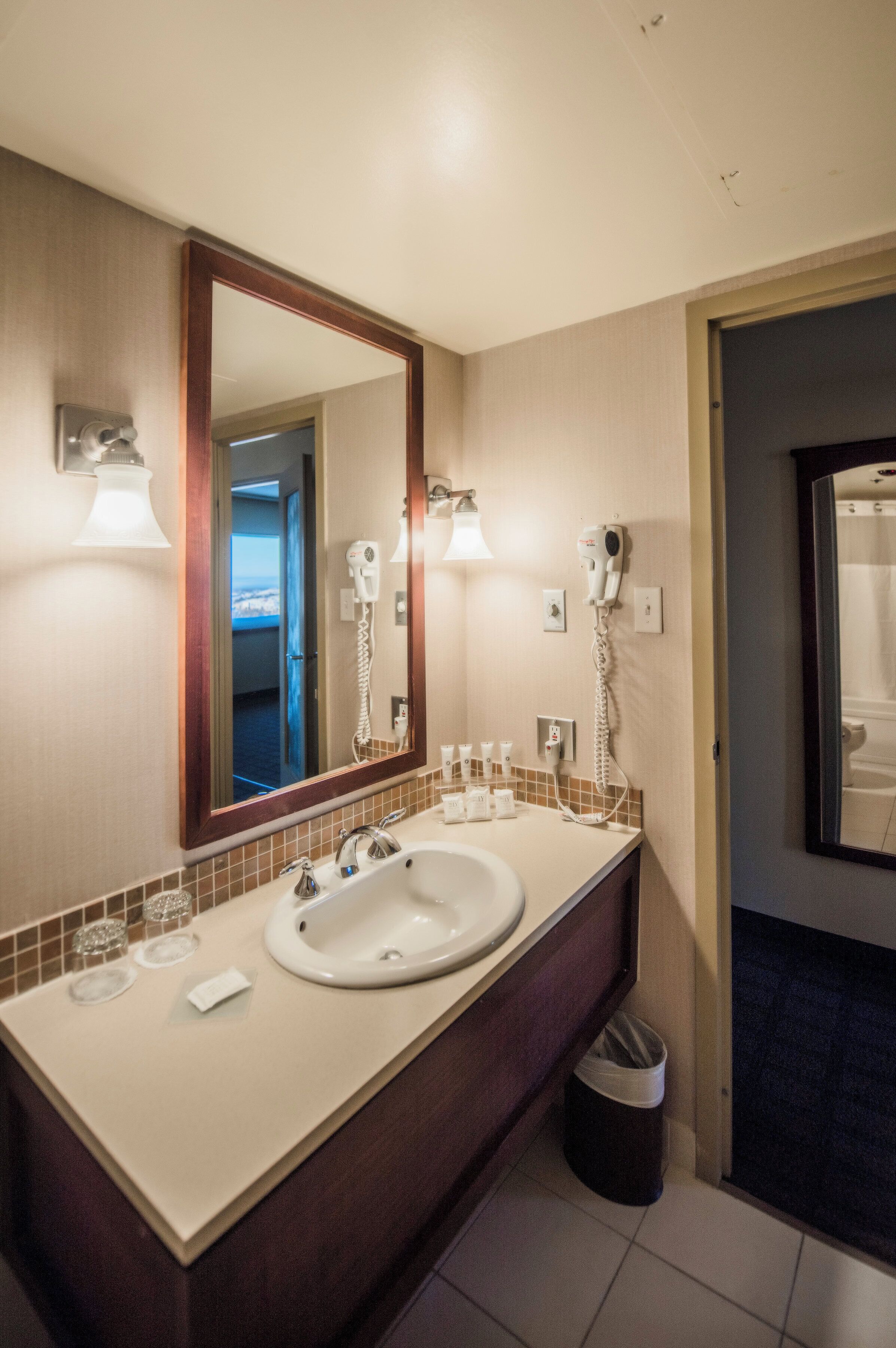 A hotel bathroom with a large mirror, two sinks, a hairdryer, and a trash bin.