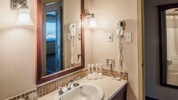 A hotel bathroom with a large mirror, two sinks, a hairdryer, and a trash bin.