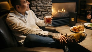 A man seated in a leather chair, holding a drink, with a stone fireplace and a plate of food in the background. - The Explorer Hotel Yellowknife (Yellowknife)
