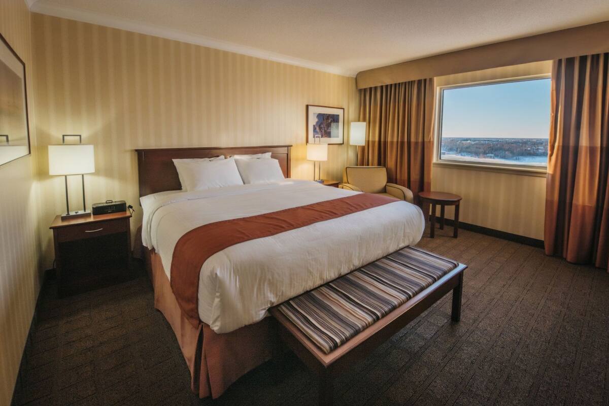 A hotel room with a large bed, a bench, a desk, a chair, a window with curtains, and a framed picture on the wall.