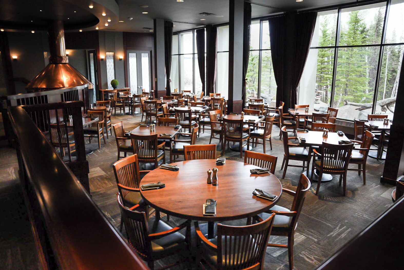 Un restaurant spacieux avec de nombreuses tables et chaises rondes, de grandes fenêtres donnant sur la forêt et une cheminée imposante de style industriel.