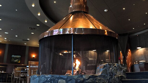 A circular stone fireplace with a metal chimney, surrounded by a stone wall and a seating area with a patterned rug. - The Explorer Hotel Yellowknife (Yellowknife)