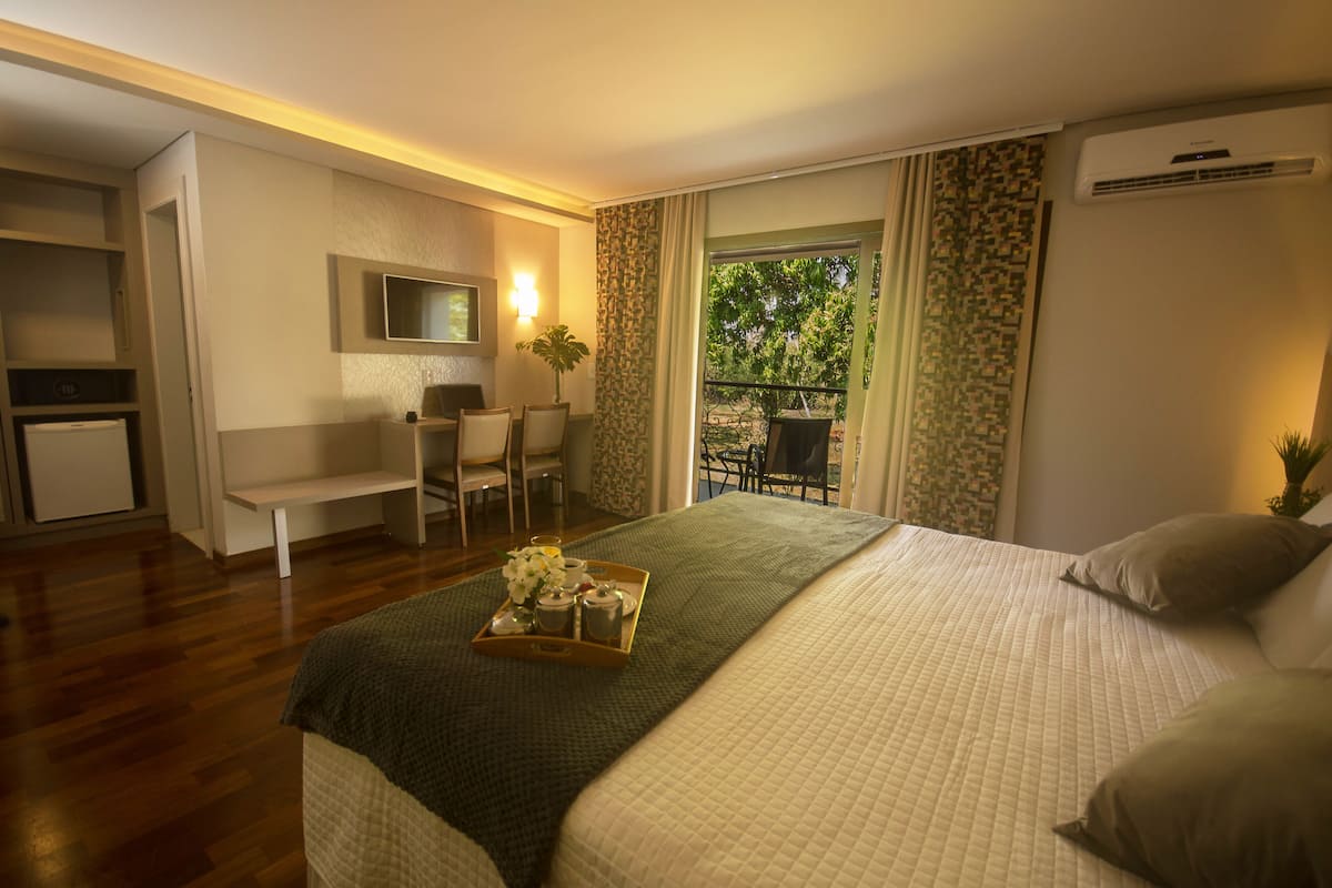 Ein Hotelzimmer mit Bett, Schreibtisch, Stuhl, einem kleinen Tisch mit Tablett und einem Balkon mit Blick auf Bäume.