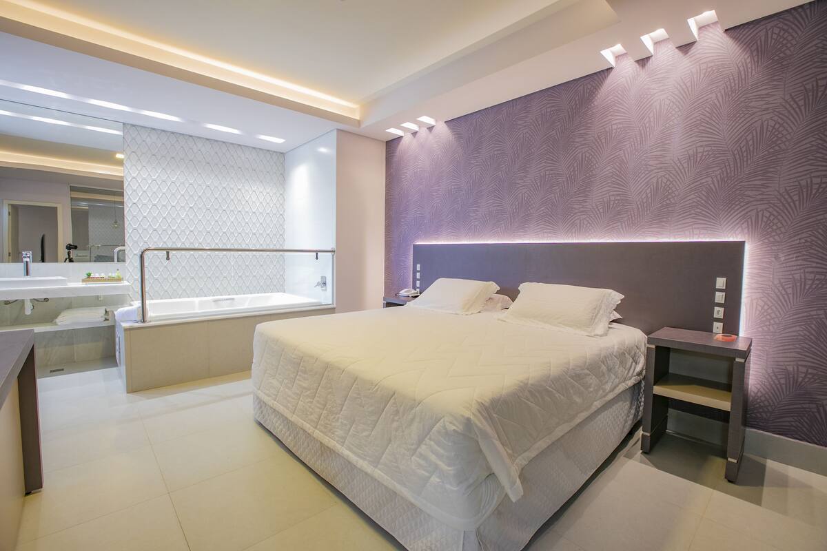 Une chambre moderne avec un grand lit, une table de chevet, une étagère intégrée et une salle de bain dotée d’une baignoire et d’un meuble-lavabo.