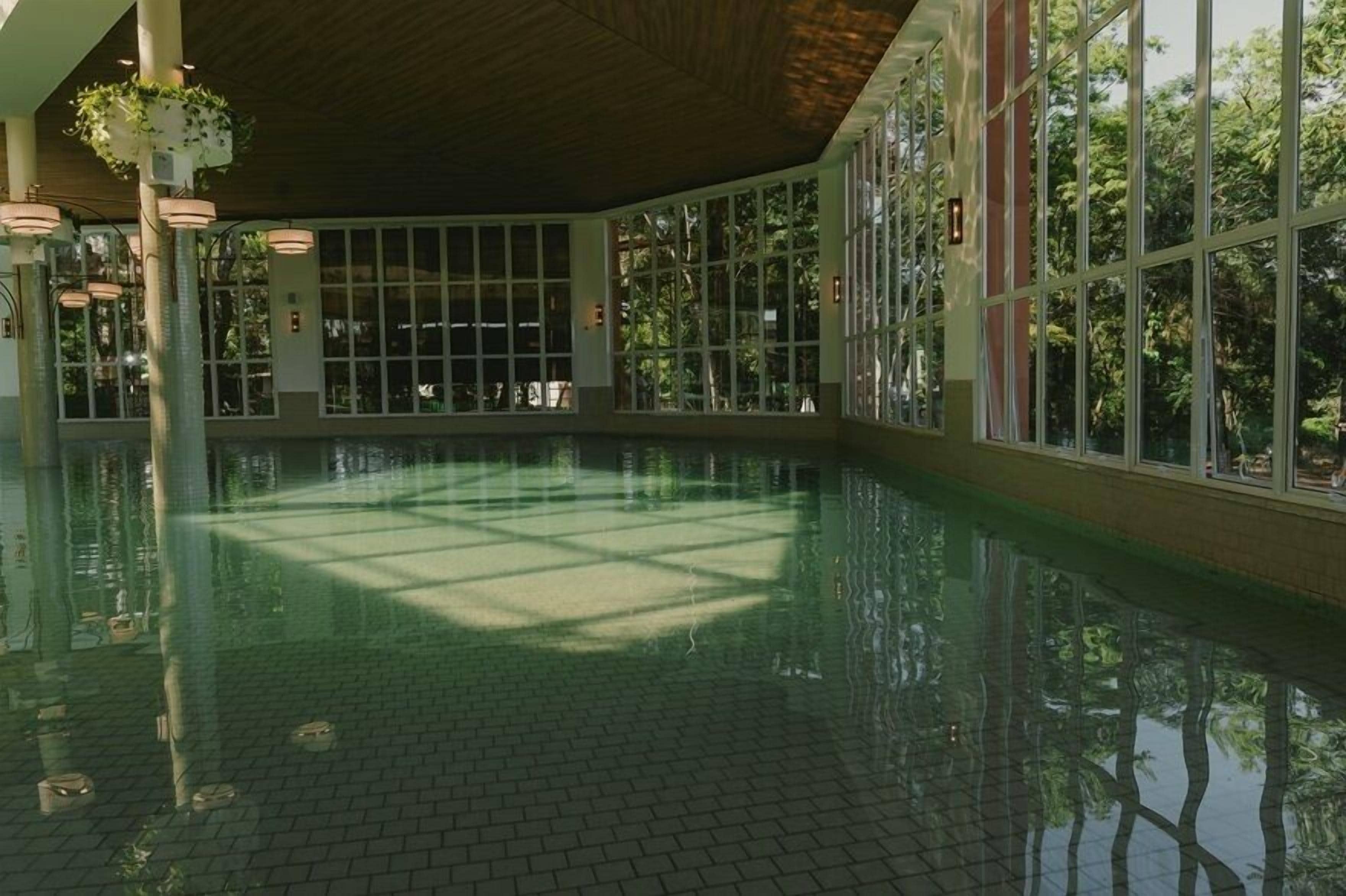 A large indoor swimming pool with clear glass walls, a tiled floor, and hanging plants.