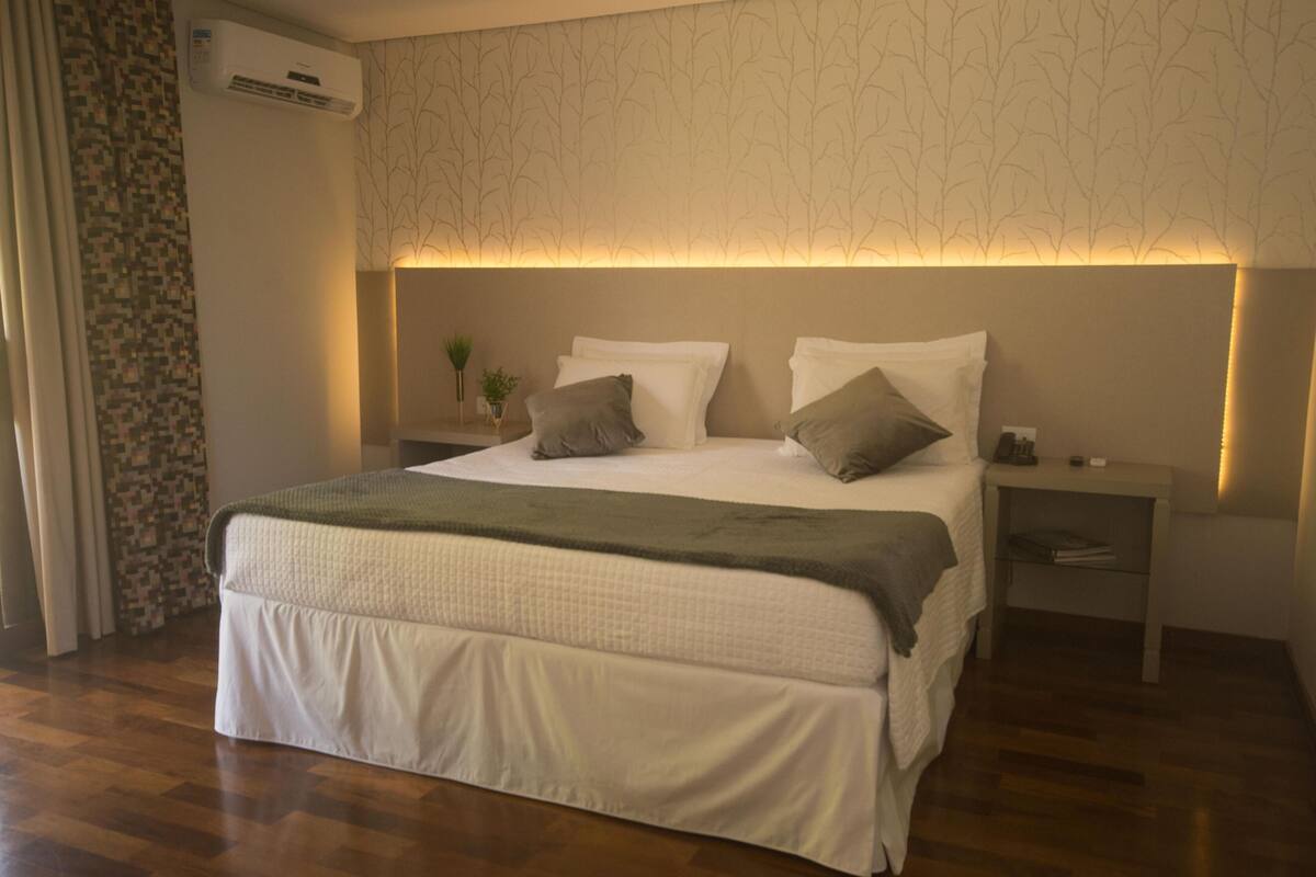 A neatly made bed with a green throw blanket, a bedside table with a lamp, and a wall-mounted air conditioner.